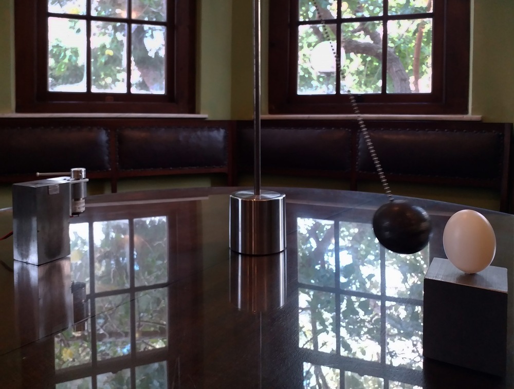 פרט מתוך עבודתה של ליטל רובינשטיין ובה נראה כדור עגול כהה קשור בחוט מתכת למבנה דמוי מטוטלת, מתנודד בעוצמה כלפי ביצה המונחת על פדסטל קטן על שולחן האוכל של ביאליק