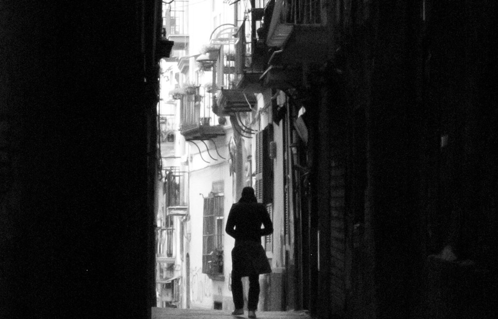 צילום בשחור ולבן בסגנון פילם נואר של גבר הנצפה מגבו פוסע לבדו ברחוב צר בין שני בניינים מוצלים ואלומת אור מאירה את דרכו